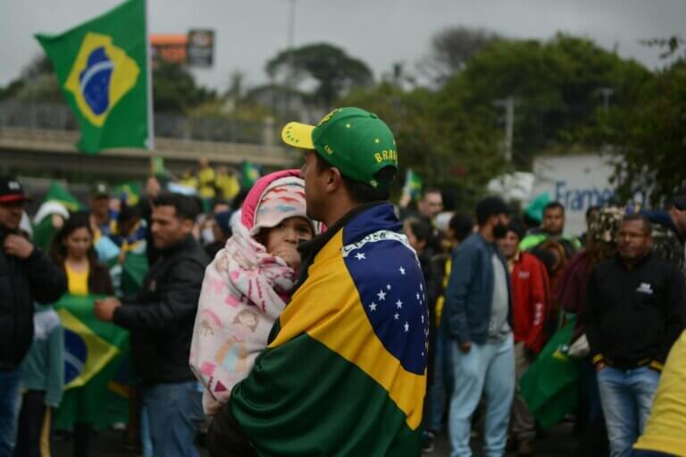 Father and child wrapped in Brazil flag at an outdoor gathering advocating democracy.